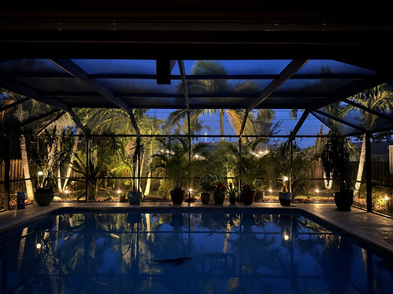 Screened pool cage at night with low-voltage landscape lighting uplighting palms and potted plants in Cape Coral